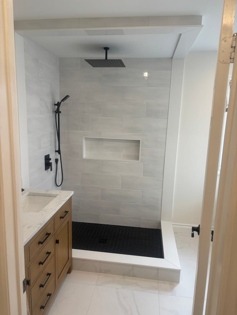 Modern bathroom with walk-in shower featuring gray tile walls, rainfall showerhead, wooden vanity with white countertop, and black shower door