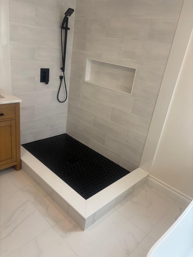 Modern bathroom shower with white tile walls, black rainfall showerhead, white frame base with black rubber floor mat, and recessed shelf